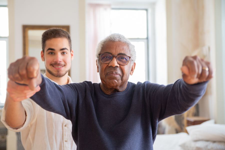 A caregiver and an older man smiling together while doing a light stretching exercise at home