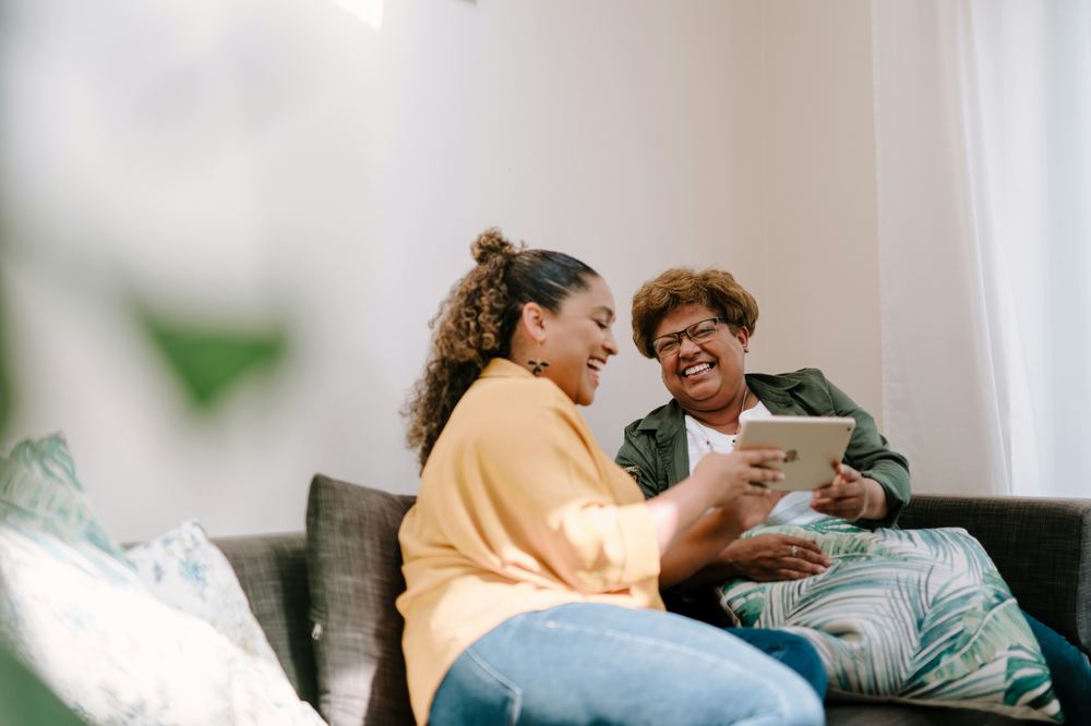 Two women laughing together on a couch while looking at a tablet