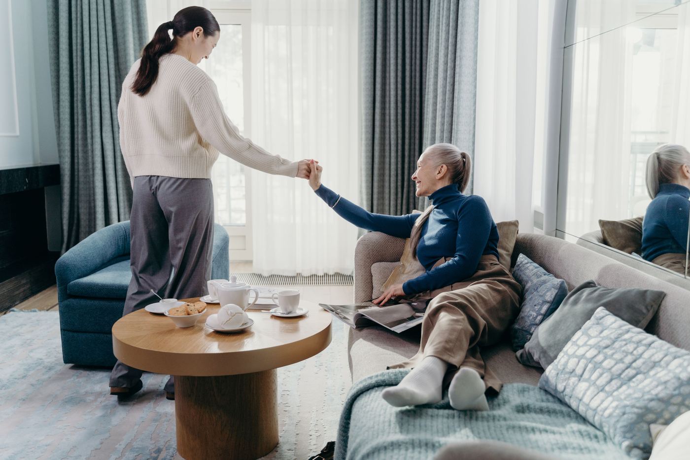 A caregiver helping an older woman up from her sofa in a bright, comfortable living room