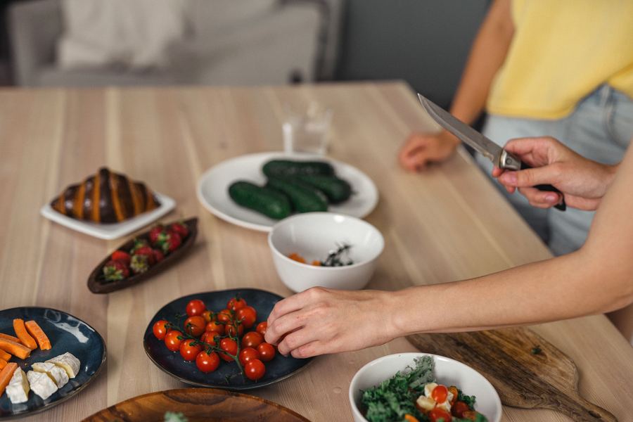 Fresh vegetables, fruit, and prepared dishes laid out on a kitchen table while someone slices ingredients