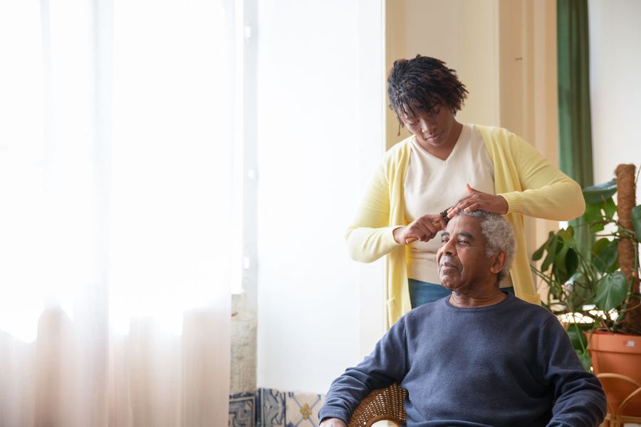 A caregiver helping an older man with his hair and grooming near a bright window